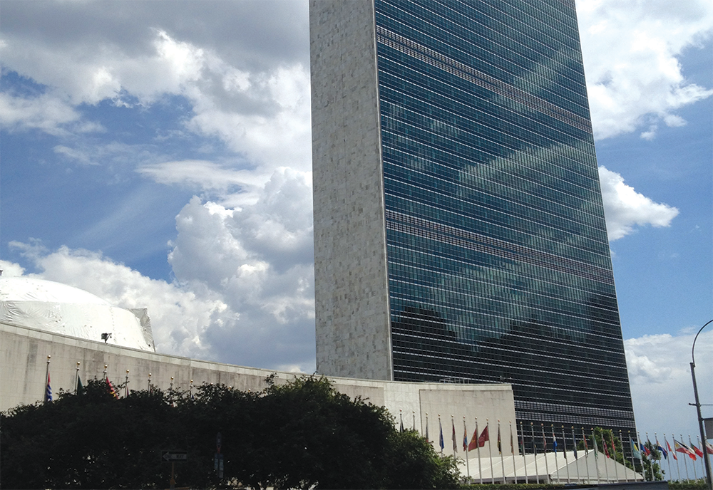United Nations Secretariat Building seen against a sunny sky with little cloud coverage and all UN member country flags in the foreground.