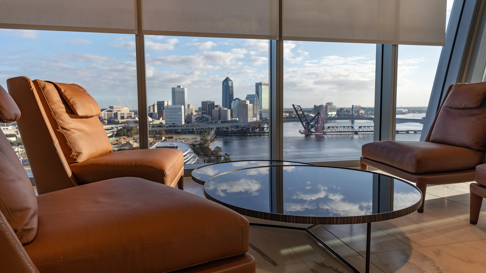 Lounge area with four chairs surrounding a table in the foreground and open windows overlooking the Jacksonville skyline in the background.